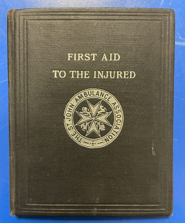 A small black book lies closed and flat on a blue metal table. The front cover is the part of the book that is visible, displaying the title debossed into the cover in white letters which read ‘First Aid to the Injured’. Below this is the emblem of St John Ambulance also debossed into the cover in white. It is an eight-pointed cross, with the words ‘The St John Ambulance Association’ surrounding it in a circle.