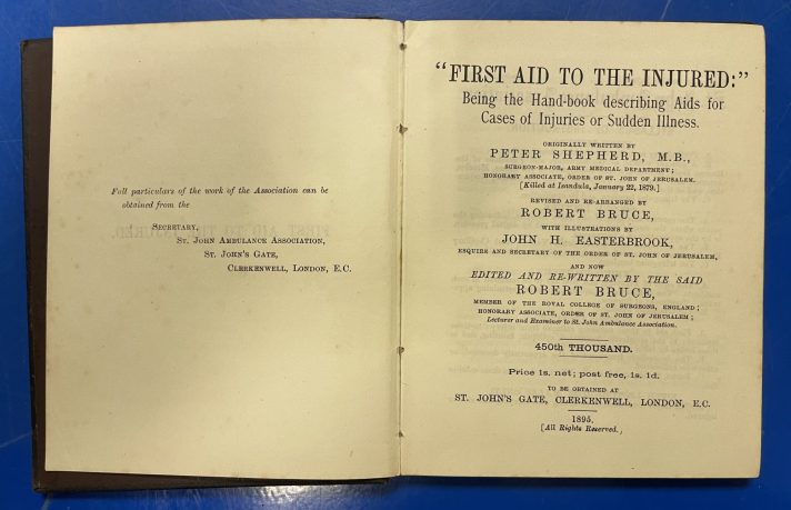 A small book lies open on a blue metal table. There is a small amount of text on the left-hand page which tells readers where they can get information on the St John Ambulance Association. The right-hand page provides the book’s title, authors, and publishing information. The text is black, and primarily in a small, serif font.