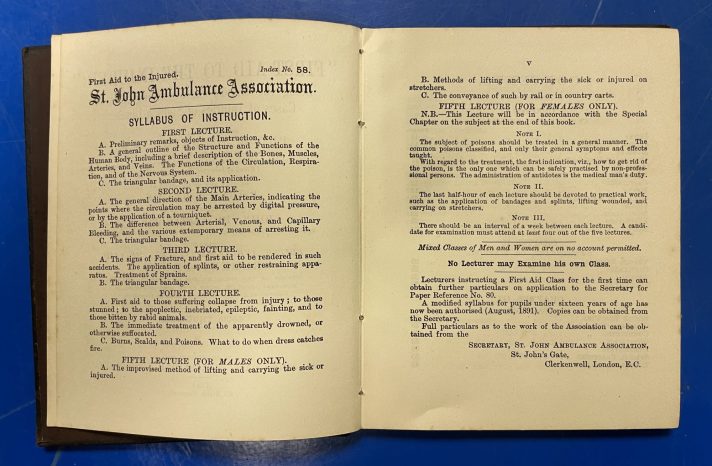 A small book lies open on a blue metal table. The pages contain lots of text in small, black, serif font. The text contains the syllabus of instruction within the manual.