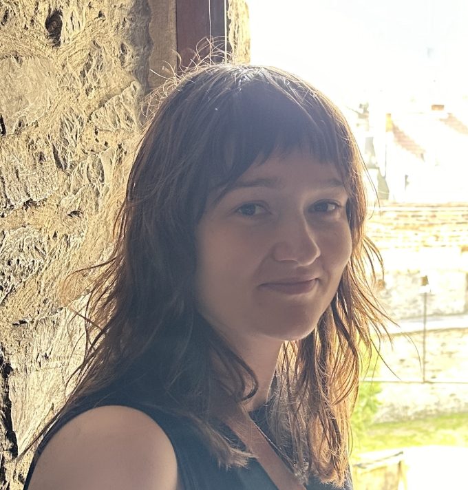 A head and shoulders colour photograph of a white woman with long brown hair and a fringe. She is standing outside with a stone wall behind her, facing slightly away from the photographer. She is wearing a dark sleeveless top and is smiling.