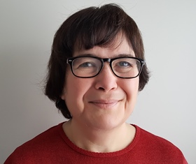 A head and shoulders colour photograph of a white woman with short brown hair, wearing a red top, and eye glasses with a dark frame. She is photographed in front of a white wall.