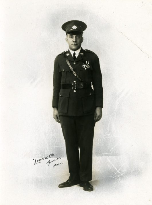 A black and white full-length photograph of a white man wearing uniform. He is wearing dark trousers and a dark jacket, with medals pinned to his left breast. He has a dark belt around his waist, and is wearing a dark cross-body bag from his right-hand shoulder. A white shirt with a dark tie is visible under his jacket. He is also wearing a dark peaked cap with a light coloured badge in the centre.