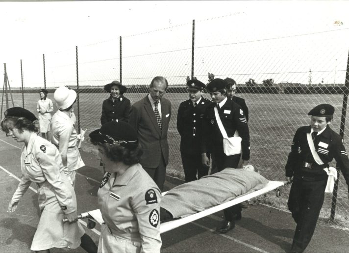 Four St John Ambulance Cadets dressed in their Cadet uniform are carrying a patient on a stretcher on a sports field, who is covered with a blanket. Two female Cadets are at the foot of the stretcher, and two male Cadets are at the head of the stretcher. Three St John Ambulance adult members in adult uniform, three civilians, and Prince Philip are standing and observing.