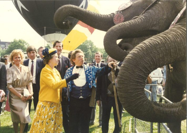 Queen Elizabeth II, dressed in a yellow dress, yellow jacket, and yellow hat, is standing next to a man dressed in a blue circus ringleader’s outfit with a white shirt and dark bow tie, and admiring three elephants. In the background is a crowd of civilians observing, and behind that, two hot air balloons are visible.