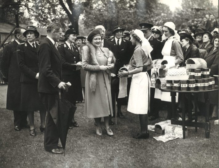 Queen Elizabeth the Queen mother is standing in the middle of a group of people. She is wearing a light coloured long coat and hat, and an animal skin scarf. She is talking to a woman, dressed in the uniform of a St John Ambulance Brigade Nursing member. Behind them is a group of men and women. Some of these people are dressed in the uniform of St John Ambulance Brigade members, and others are in civilian dress.