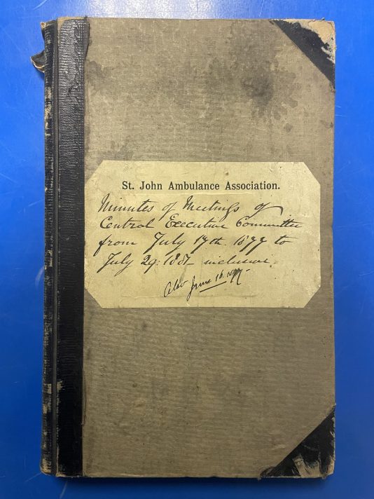 Two big scrapbooks lie on top of a blue metal table, one on top of the other. The books are large bound items made of a dark beige covered board with a dark leather-covered spine and corners. On the front of the top book is a white label with text which reads ‘St. John Ambulance Association. Minutes of Meetings of Central Executive Committe from July 17th 1877 to July 19 1881 – inclusive. Also June 16 1877’.