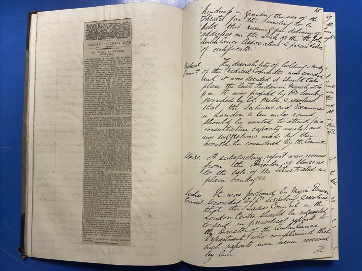 A large scrapbook lies open on a blue metal table. The right-hand page contains handwritten minutes from a meeting, written in black ink. The left-hand page has a newspaper article about St John pasted in.