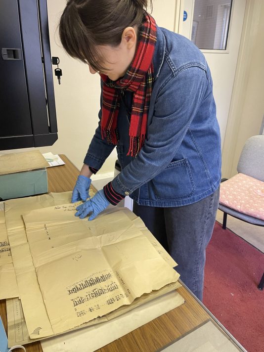 A white woman with dark hair tied into a ponytail, wearing a red tartan scarf, blue denim jacket and grey trousers is standing next to a table. She is looking at paperwork which is spread out on top of the table.