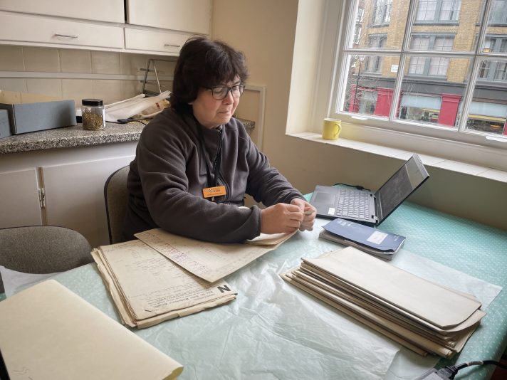 A white woman in glasses with dark hair wearing a dark jumper is sitting at a table covered in a green cloth, next to a window. On the table in front of her is a laptop, and piles of documents. She is leafing through the documents.