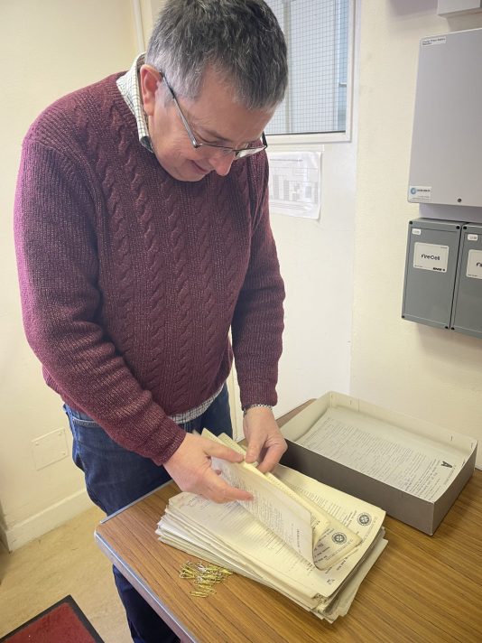 A man with grey hair and glasses is standing over a brown table looking at some documents. He is wearing a red jumper with a pale shirt underneath, and blue denim jeans. On the table in front of him is an open grey cardboard box, with a pile of documents next to it. The man is leafing through the documents. 