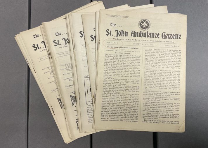 A set of around 10 small off-white booklets are fanned out on top of grey cardboard. The booklets are newsletters called The St. John Ambulance Gazette, and were published by St John Ambulance in Australia. 